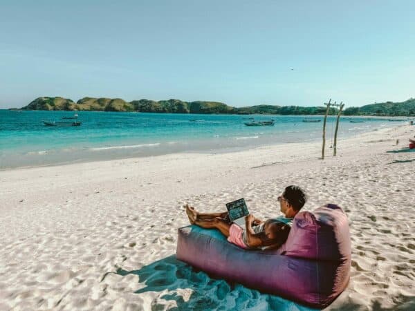 Digital nomade jobber på laptop fra en tropisk strand med turkist hav i bakgrunnen.