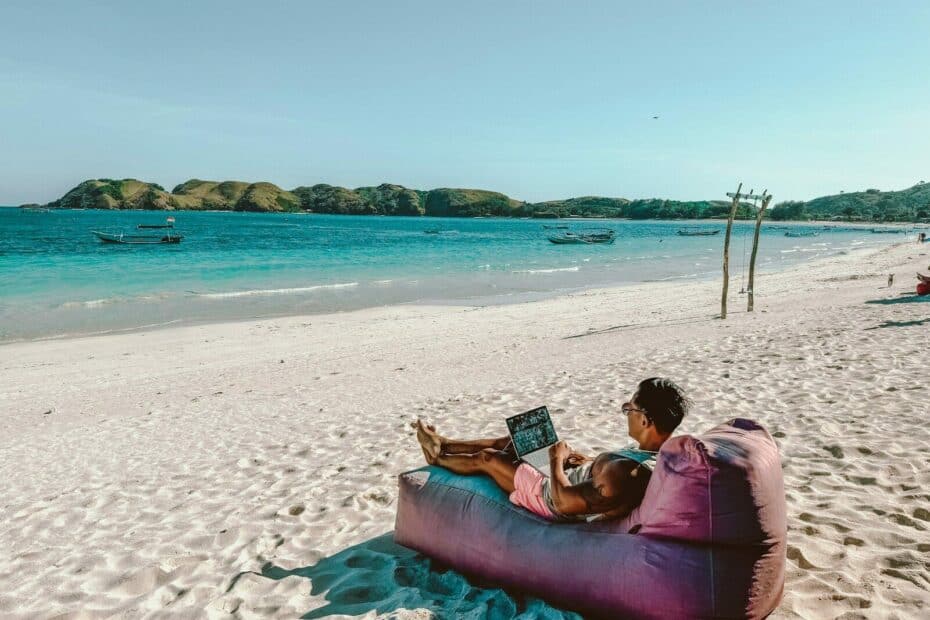 Digital nomade jobber på laptop fra en tropisk strand med turkist hav i bakgrunnen.