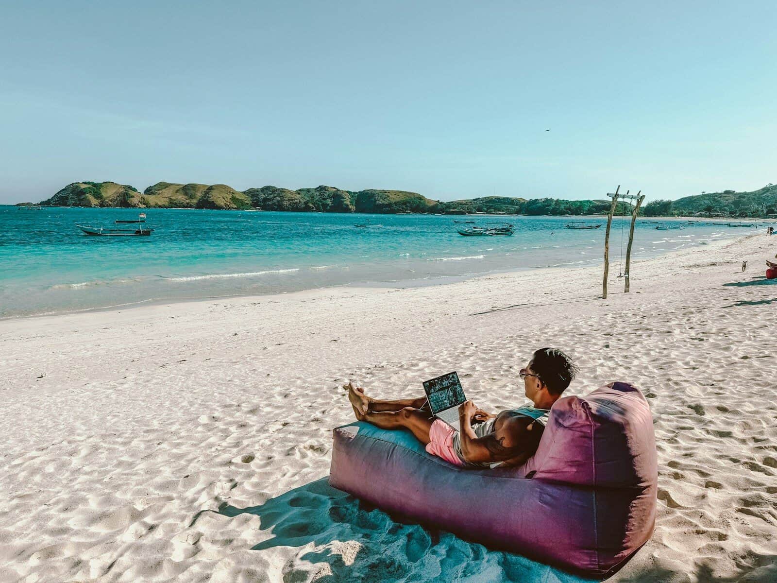Digital nomade jobber på laptop fra en tropisk strand med turkist hav i bakgrunnen.