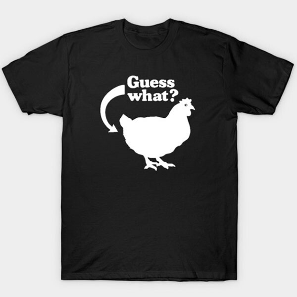 Svart T‑skjorte med hvit tekst «Guess What?» og en enkel hønefigur med pil som peker på fuglens bakende.