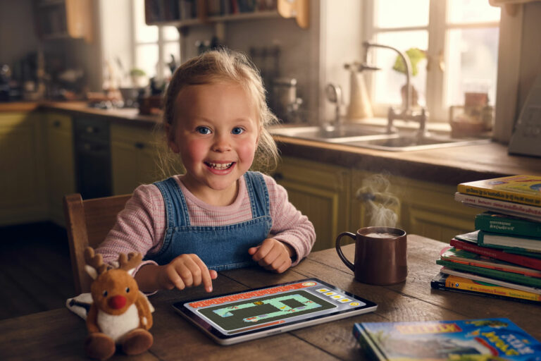 Smilende liten jente bruker kode-app på nettbrett ved kjøkkenbordet, omgitt av bøker, kakao og en kosedyr.